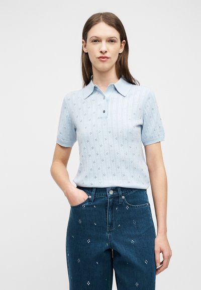 Γυναίκα με ίσια καστανά μαλλιά φοράει ανοιχτό μπλε πλεκτό πόλο με κοντά μανίκια και ψηλόμεσα σκούρα μπλε τζιν με μικρά λευκά κεντημένα σχέδια.