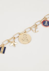 Bracelet charm plaqué or avec une étoile, une ancre, un médaillon circulaire et une étiquette rayée. Maillons épais avec une finition brillante et des éléments de design variés.