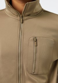 Beige zip-up jacka med hög krage, som har en vertikal dragkedjeficka på vänster bröst och textilstruktur. Dragkedjor i matchande färg.