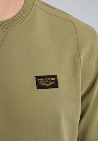 Felpa verde oliva con scollo a coste e un'etichetta rettangolare nera con testo e logo giallo sul petto.