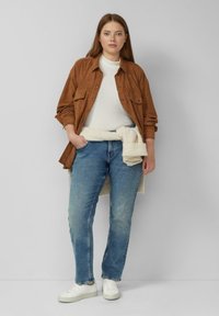 Brązowa koszula zapinana na guziki na białym prążkowanym swetrze, jasnoniebieskie dżinsy i białe trampki. Model stoi z rękami w kieszeniach.