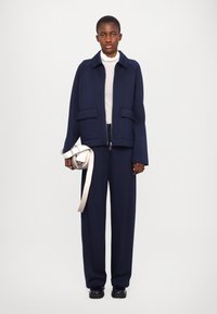 Navy blue jacket with wide sleeves and two front pockets, paired with matching trousers and a light gray turtleneck, holding a cream crossbody bag.