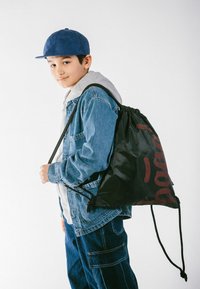 Zaino con coulisse nero con motivo rosso, realizzato in materiale leggero. Indossato da un bambino con una giacca di jeans blu e un felpa grigia.