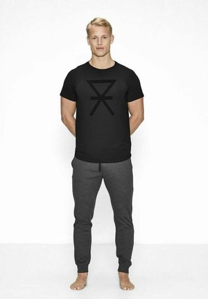 Sort t-shirt med et vandret stribet grafik på brystet, parret med grå sweatpants. Model står barfodet med en neutral holdning.