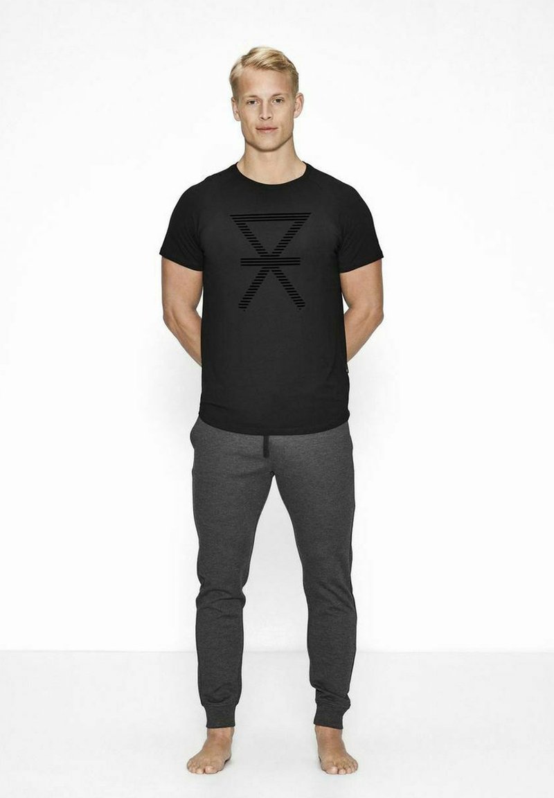 Sort t-shirt med et vandret stribet grafik på brystet, parret med grå sweatpants. Model står barfodet med en neutral holdning.