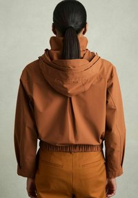 Giacca impermeabile marrone con cappuccio, dotata di una vita elasticizzata e maniche lunghe, abbinata a pantaloni marroni coordinati.