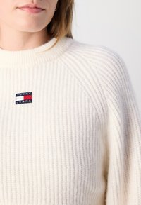 White ribbed knit sweater featuring a round neckline and a small embroidered logo patch in red, white, and blue on the chest.