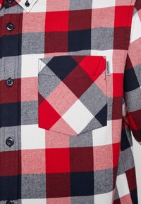 Camisa a cuadros en tonos rojo, blanco y azul marino, con cuello abotonado y bolsillo en el pecho. La tela tiene un aspecto texturizado y tejido.