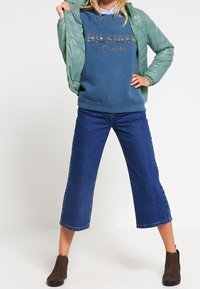 Person i grön pufferjacka över en blå "Hilfiger Denim" sweatshirt, vida avklippta jeans och bruna ankelstövlar, står med benen isär.