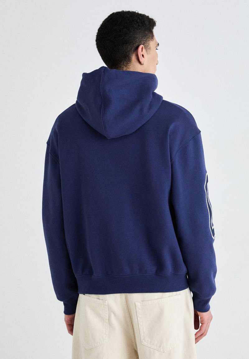 Dunkelblaues Hoodie aus weichem Stoff, mit großer Kapuze, gerippten Bündchen und einem Logo auf dem linken Ärmel. Lockere Passform, minimales Design.