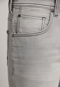 Los jeans de mezclilla gris presentan un corte ajustado con costuras visibles a lo largo de los bolsillos, dos bolsillos frontales y una textura sutil.