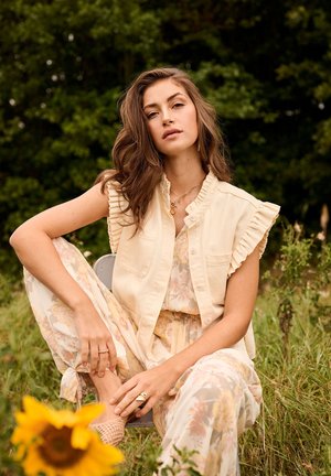 Junge Frau mit langen braunen Haaren, die geblümte Hose und eine ärmellose, geraffte Weste trägt, sitzt draußen zwischen Grün und einer Sonnenblume.