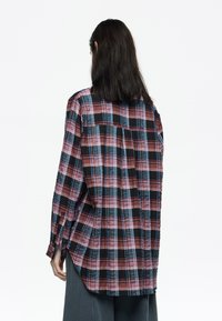 Camisa de cuadros en tonalidades oscuras de rojo, azul y negro, con mangas largas y un corte relajado que incluye un pliegue en la parte posterior para facilitar el movimiento.