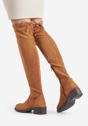 Žena nosící béžové semišové kozačky po kolena s nízkými širokými podpatky a zipy na kotnících, doplněné o bílou žebrovanou sukni.