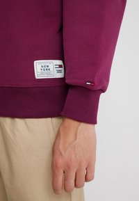 Vínově červená mikina s žebrovanými manžetami a lemem, která nese bílou štítek s nápisy "NEW YORK" a "TOMMY JEANS". Malý logo prvek u manžety.