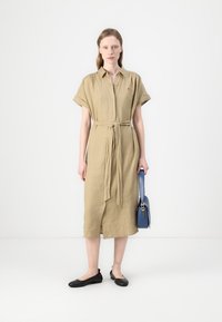 Rochie din in khaki, cu mâneci scurte rulate, închidere cu nasturi în față și talie legată. Asortată cu pantofi negri și o geantă albastră.