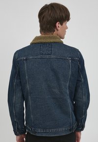 Tumši zila džinsa jaka ar mīkstu, brūnganu mākslīgās kažokādas apkakli. Aprīkota ar pogām aizdarāmu priekšu un aprocēm, kā arī dubultšuvēm gar šuvēm.