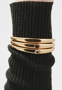 Brazaletes de oro en forma de bangle con tres bandas lisas y redondeadas, que presentan una piedra preciosa rosa y una piedra preciosa clara, llevados sobre una manga negra texturizada.