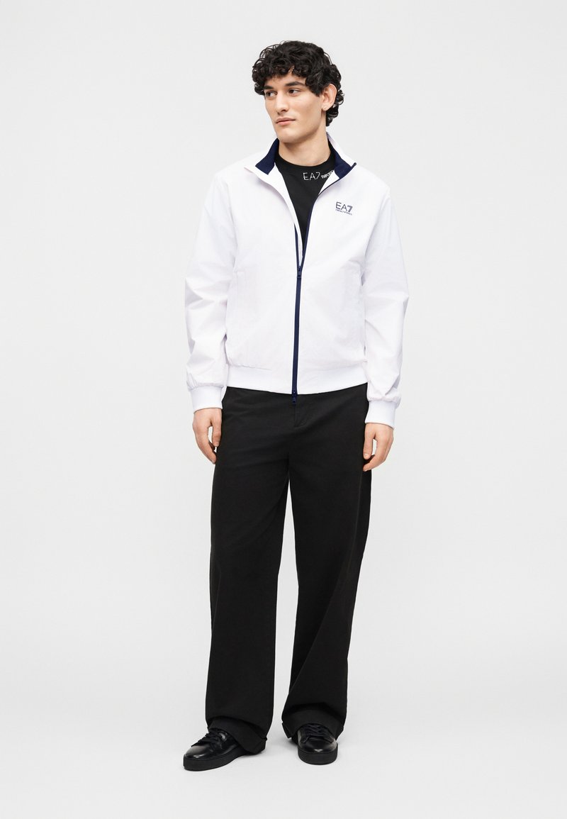 Νέος άνδρας που φοράει λευκό τζάκετ με φερμουάρ πάνω από μαύρο πουκάμισο EA7, μαύρο παντελόνι με φαρδιά πόδια και μαύρα αθλητικά παπούτσια, στέκεται μπροστά σε λευκό φόντο.