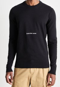 Svart tröja med långa ärmar i mjukt stickat material. Har ribbstickad halsringning och muddar. "Calvin Klein Jeans"-logotyp i vitt.