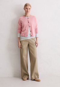 Cardigan rose clair à col rond et boutons marron, superposé sur une chemise à rayures bleues. Pantalon large beige et chaussures beiges.