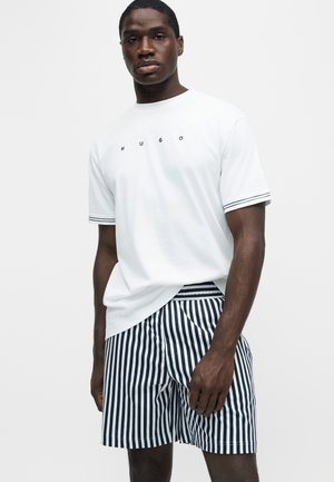 Μαύρος άνδρας φοράει λευκό T-shirt με κείμενο "HUGO" και σκούρο μπλε και λευκές κάθετες ριγέ βερμούδες, στέκεται μπροστά σε απλό λευκό φόντο.