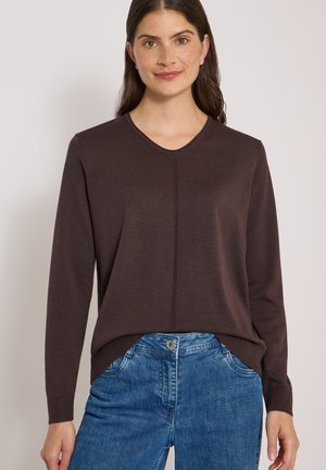 Kvinde iført en mørkebrun langærmet sweater og blå jeans, smilende mod en ensfarvet lys baggrund.