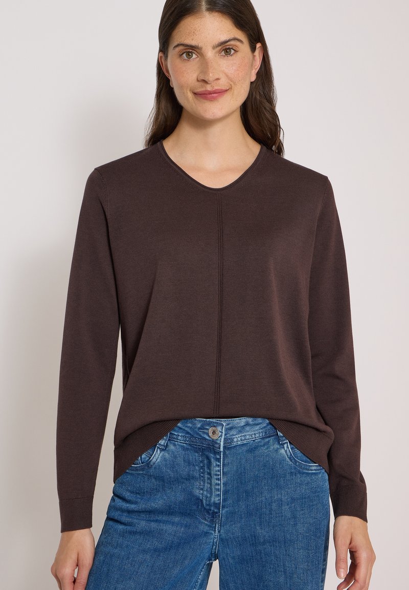 Femme portant un pull marron foncé à manches longues et un jean bleu, souriant devant un fond clair uni.