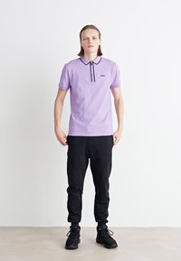 Fialová polo košile s černým límcem a lemováním, spárovaná s černými jogger kalhotami. Oblečení má volný střih a neformální styl.