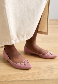 Ljust rosa mocka ballerinaskor med spetsig tå, prydda med en guldbåge vid framfoten och en låg, texturerad sula.