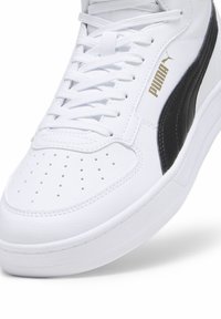 Baskets blanches Puma avec bande latérale noire, bout perforé, lacets blancs et logo Puma doré sur le côté.