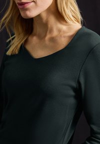 Černý top s výstřihem do V vyrobený z hladké strukturované tkaniny. Má dlouhé rukávy a těsný střih kolem výstřihu, což zvýrazňuje oblast ramen.