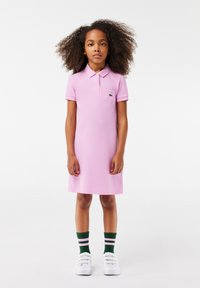 Vestido polo de algodón rosa claro con mangas cortas, un cuello y un bolsillo en el pecho; combinado con calcetas a rayas verdes y rosas y zapatos blancos.