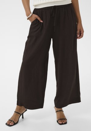 Mujer con pantalones anchos negros de pierna ancha y cintura con cordón, una mano en el bolsillo, combinados con sandalias negras de tacón.