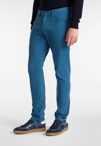 Blue cotton blend jeans with a slim fit, five-pocket design, and front button closure, paired with dark sneakers featuring suede and rubber accents.