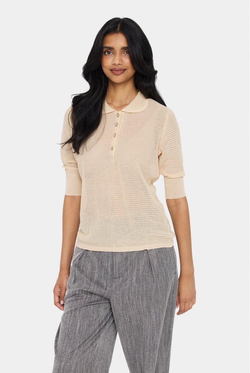 Polo shirt beige in maglia con maniche corte, chiusura a bottoni e motivo testurizzato; indossato con pantaloni grigi a pieghe.