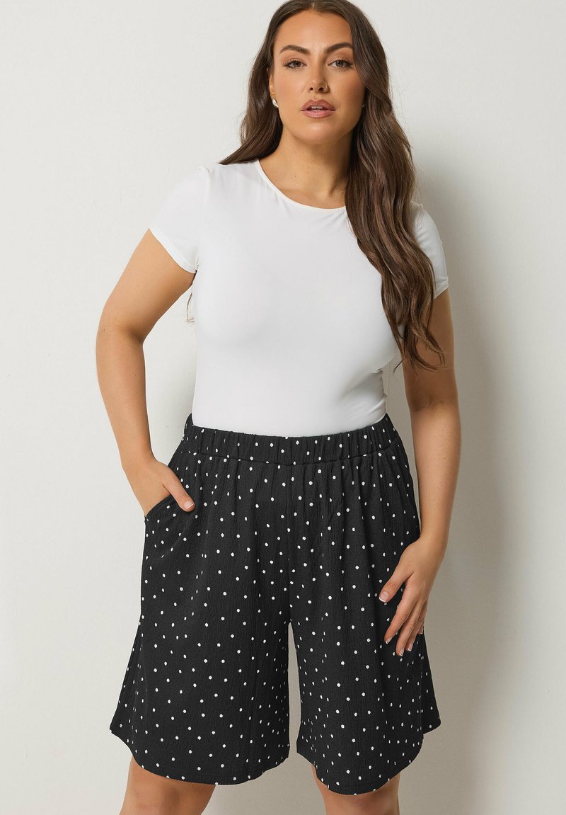 Vrouw met lang bruin haar, gekleed in een nauwsluitend wit T-shirt met korte mouwen en zwarte knielange short met witte stippen, staand met handen in de zakken.