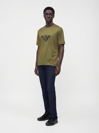 Мъж, облечен в тениска с лого на Emporio Armani в маслинено зелено, тъмносини дънки и черни маратонки, стои пред бял фон.