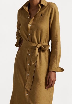 Mujer con un vestido largo de manga larga color mostaza marrón con botones, cinturón de tela atado a la cintura y un pequeño logo bordado en el pecho.