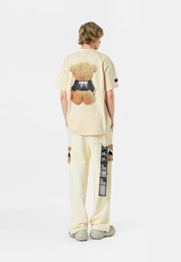 Camiseta beige de gran tamaño con un gráfico de oso difuso en la parte posterior, combinada con pantalones crema adornados con aplicaciones de osos y una etiqueta.