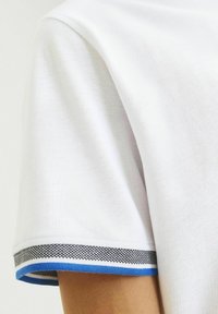 Weißes Polohemd aus strukturiertem Stoff. Der Kragen ist mit einem blau-schwarzen gestreiften Rand entlang der Kanten versehen.