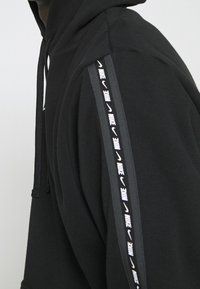 Sudadera negra con logo de Nike y un patrón de swoosh que baja por la manga izquierda, usada por una persona que está ligeramente de lado.