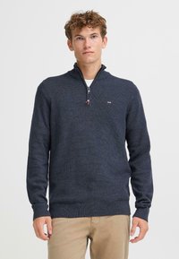 Sweater azul marino con cremallera y un tejido texturizado, con cuello alzado. Presenta mangas largas y un pequeño logo de la marca en el pecho.