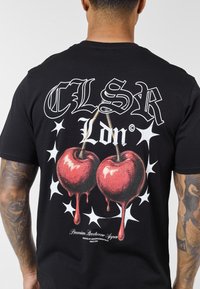 Μαύρο μπλουζάκι με γραφικό δύο κόκκινων κερασιών με λεπτομέρειες που στάζουν και στοιχεία αστεριών, με το κείμενο «CLSR Ldn.» Το υλικό φαίνεται βαμβακερό.