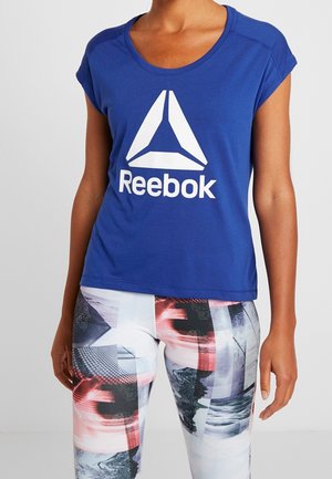 Osoba nosząca niebieską koszulkę Reebok w połączeniu z legginsami o abstrakcyjnym wzorze w kolorach białym, czarnym i czerwonym, stojąca na jednolitym tle.