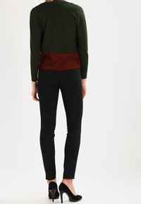 Zielony krótki kardigan nałożony na rdzawą bluzkę, w zestawieniu z czarnymi dopasowanymi spodniami i czarnymi szpilkami. Gładki materiał, minimalistyczny design.
