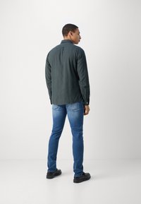 Dark green button-up shirt with long sleeves, a back yoke, and a relaxed fit; paired with blue slim-fit jeans and black shoes.