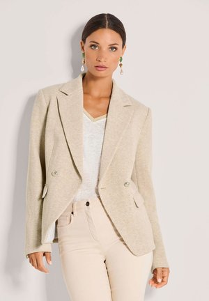 Beiger Fischgräten-Blazer mit doppelt geknöpften Knöpfen, tailliertem Schnitt und schrägen Taschen, kombiniert mit einem weißen V-Ausschnitt-Oberteil und hellen Hosen.