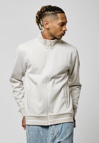 Chaqueta cremosa con cremallera y un patrón de cuadrícula texturizado, cuello alto y bolsillos laterales, combinada con jeans azul claro ligeramente desgastados.