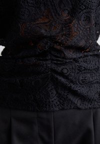 Tissu en dentelle noire avec des motifs floraux et circulaires complexes superposés sur un pantalon noir, mettant en valeur des détails froncés au centre.